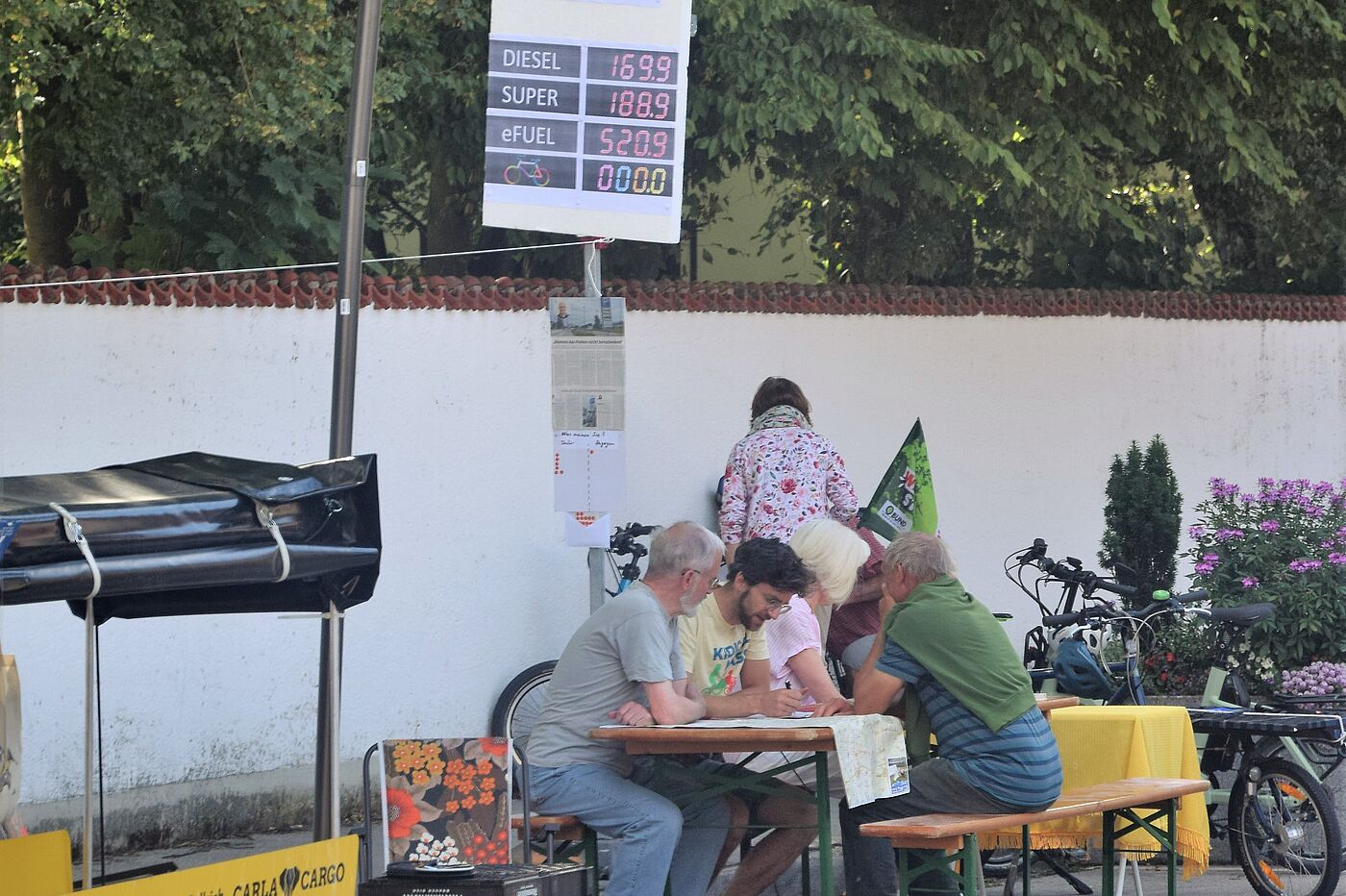 Tankstelle Personen auf Biertischgarnitur, davor eine Tankstellenpreistafel