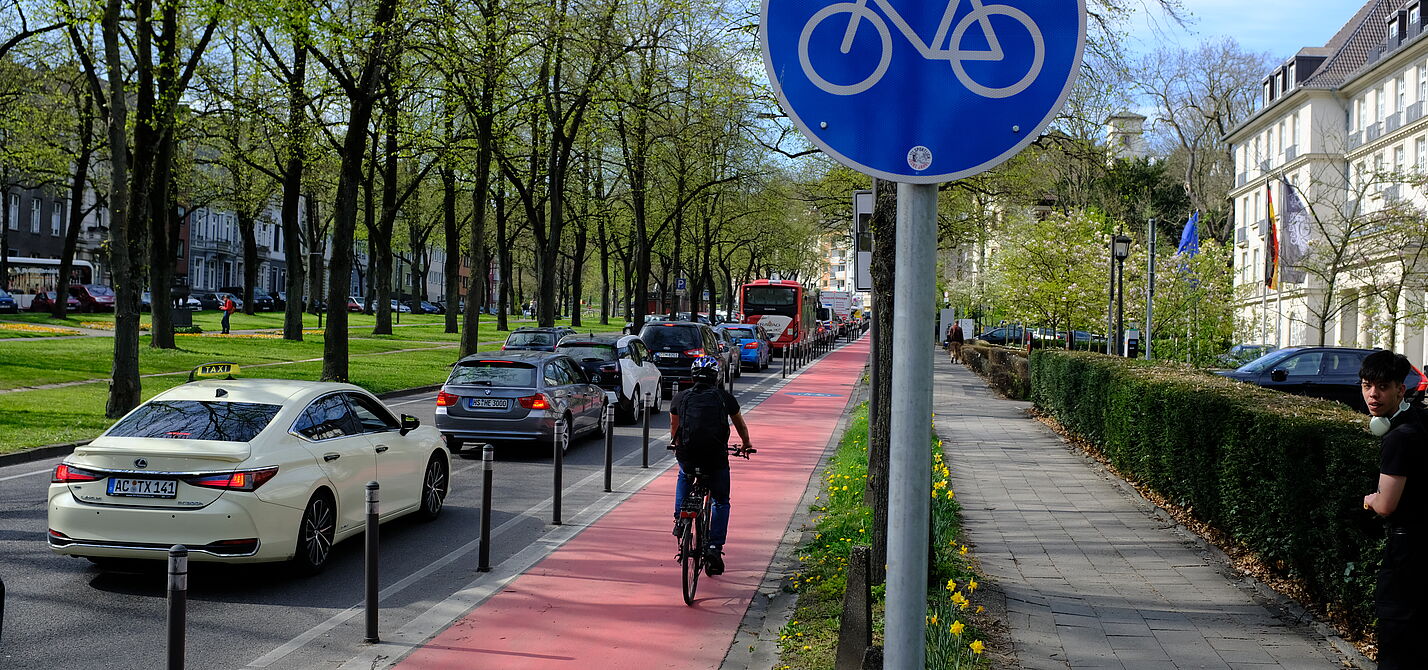 Ein geschützter Radfahrstreifen mit Roteinfärbung. Radwegschild und einer Radfahrerin. 