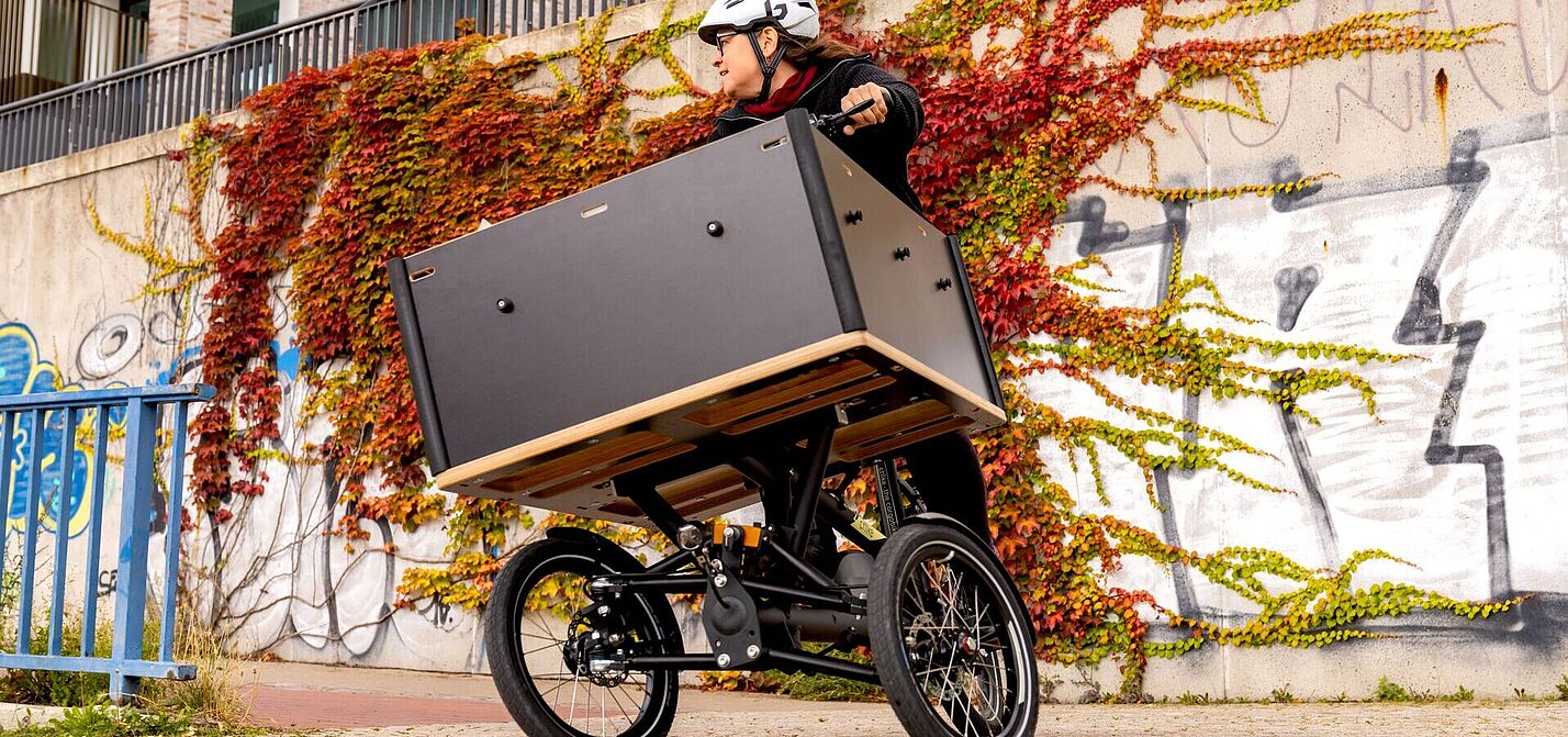 Person mit Helm fährt ein dreirädriges Lastenrad mit großer Transportbox vor einer bewachsenen Wand.