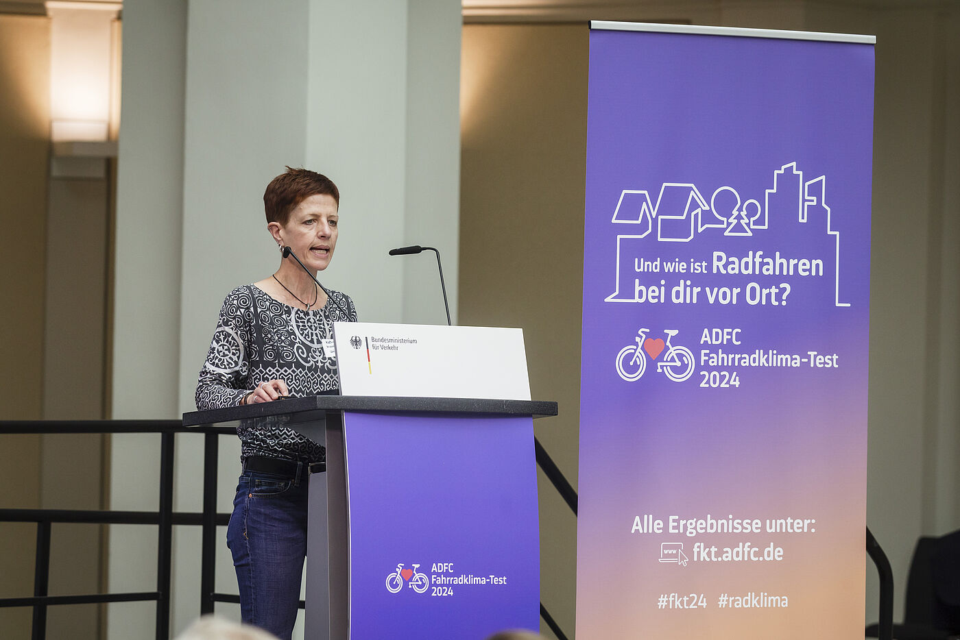 Rednerin am Podium Prof. Dr. Katharina Manderscheid, Frau mit kurzen braunen Haaren in gemustertem Oberteil, steht am Rednerpult mit Mikrofon vor lila ADFC-Banner mit Aufschrift „Und wie ist Radfahren bei dir vor Ort? ADFC Fahrradklima-Test 2024"